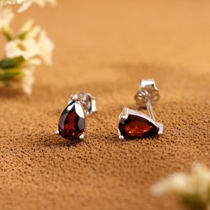 Könnte beinhalten: Ein Paar Ohrstecker aus Silber mit tropfenförmigen, tiefroten Edelsteinen. Die Ohrringe sind vor einem strukturierten, braunen Hintergrund platziert, mit weißen Blumen oben links und unten rechts.