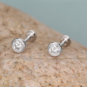 May include: A pair of small, silver stud earrings with a round, clear gemstone set in a bezel. The earrings are displayed on a textured, light brown surface against a soft blue background.