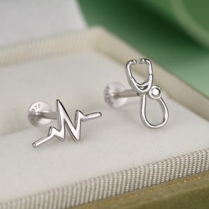 May include: A pair of silver stud earrings featuring a medical theme. One earring is shaped like a heartbeat EKG line, and the other is shaped like a stethoscope. Both earrings have a flat circular backing and are made of polished silver metal.