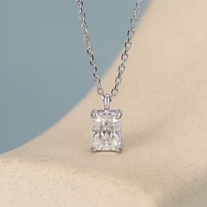 May include: A silver-toned necklace featuring a rectangular, clear, faceted gemstone pendant held by four prongs. The pendant is suspended from a delicate, thin metal chain with small, uniform oval links, creating a simple and refined jewelry piece.