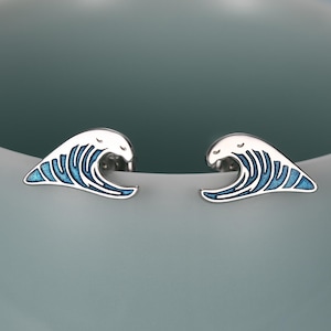 May include: A pair of silver wave-shaped stud earrings with blue enamel detailing. The earrings are set against a soft blue background, showcasing their minimalist design and ocean-inspired theme.