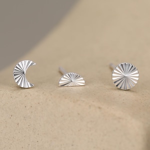 May include: Three silver stud earrings featuring a sunburst texture. The set includes one crescent moon shape, one half-circle shape, and one full circular shape. Each piece has a polished metallic finish with fine engraved lines radiating from the center.