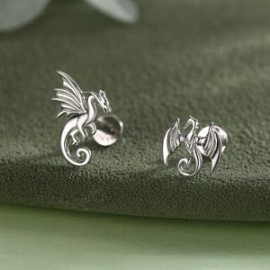 May include: Two silver dragon stud earrings. Each earring features a detailed dragon design with outstretched wings. The dragons are facing different directions, and the earrings are set against a green background.