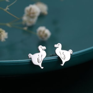 Pode incluir: Um par de brincos de prata em forma de dodô sobre um fundo turquesa.