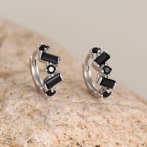 May include: A pair of silver hoop earrings with a geometric design. Each earring features a series of black rectangular and round stones. The earrings are displayed on a textured, light brown surface.