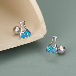 Könnte beinhalten: Ein Paar silberfarbene Ohrstecker. Jeder Ohrring hat ein kleines, blaues Design eines wissenschaftlichen Kolbens mit zwei weißen Punkten. Die Ohrringe haben einen Stecker und einen Kugelverschluss. Der Hintergrund ist hellgrün.