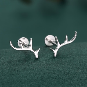 May include: A pair of silver stud earrings in the shape of deer antlers. The earrings have a polished finish and are small in size. They are displayed against a dark green backdrop.