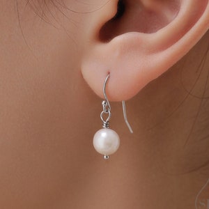May include: A single pearl earring with a silver hook is shown. The earring features a round, white pearl suspended from a silver wire. The earring is displayed next to an ear, showcasing the design and size of the jewelry.