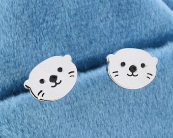 Può includere: Un paio di orecchini a perno in argento con un design di simpatica lontra. Gli orecchini sono esposti in una scatola per gioielli in velluto blu.