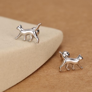 May include: Two silver cat-shaped stud earrings. The earrings are in the shape of a walking cat with a curved tail. The earrings are on a light beige surface with a brown background.