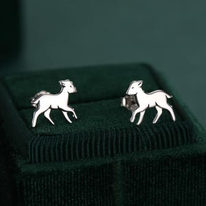 May include: A pair of silver-colored deer stud earrings. The earrings are displayed on a dark green velvet jewelry box. The deer are in a running pose. The background is a dark green color.