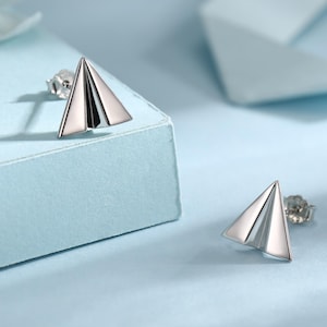 Può includere: Un paio di orecchini a bottone argentati a forma di aeroplano di carta. Gli orecchini sono esposti su una superficie azzurra. Gli orecchini sono piccoli, di forma triangolare e hanno una finitura metallica lucida. Il design è minimalista e moderno.