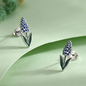 Puede incluir: Un par de pendientes de botón con un diseño de flor azul y hojas verdes. Los pendientes tienen un acabado plateado. La flor tiene un aspecto detallado y texturizado. Fondo verde claro.