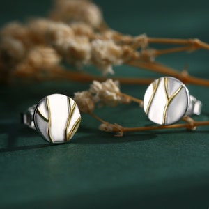 Op de afbeelding: Zilveren oorstekers met een wit emaille middenstuk en goudkleurige, takachtige accenten. De ronde oorbellen zijn geplaatst tegen een donkergroene achtergrond met gedroogde bloemen.