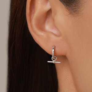 May include: A small silver-toned hoop earring featuring a dangling toggle bar charm. The earring consists of a polished circular hoop that connects to a horizontal metal bar, creating a simple and clean design that hangs from the earlobe.