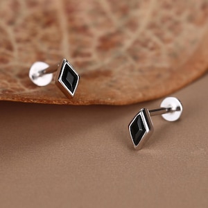 Può includere: Due orecchini a lobo color argento con una pietra nera a forma di diamante. Gli orecchini sono esposti su una superficie marrone con una foglia sullo sfondo. Gli orecchini hanno un perno e un retro piatto.