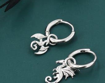 May include: Two silver hoop earrings with dragon pendants. The earrings are set against a dark teal backdrop with a black leafy branch. The dragon pendants feature detailed wings and scales.