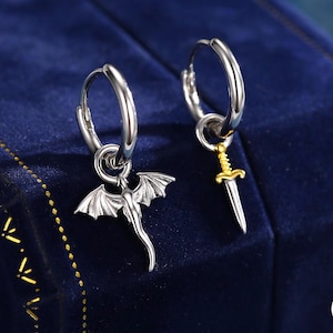 Puede incluir: Un par de pendientes de aro plateados con colgantes únicos. Uno presenta un colgante de dragón, el otro una espada dorada. Los pendientes se exhiben sobre una superficie de terciopelo azul oscuro.