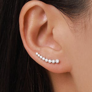 May include: A silver ear climber earring featuring a curved design with graduated, round, clear gemstones. The earring is shown on an ear, with the gemstones ascending in size.