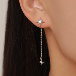 May include: A silver-toned earring featuring a starburst-shaped stud with a central clear stone. A delicate chain hangs from the stud, ending in a smaller matching starburst charm. The design is crafted from polished metal with a bright finish.