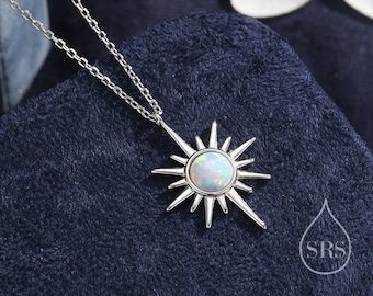May include: A silver necklace with a sunburst pendant featuring a round, iridescent opal stone. The pendant has pointed rays extending outwards. The necklace is displayed on a dark blue, textured surface, with a white flower in the background.