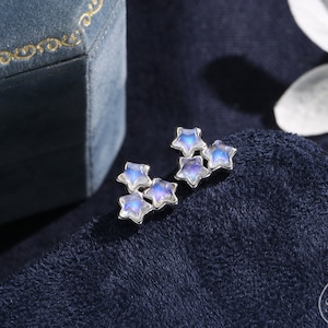 May include: A pair of silver earrings featuring a cluster of five iridescent blue star-shaped stones. The earrings are displayed on a dark blue fabric background.