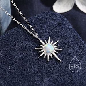 May include: A silver necklace with a sunburst pendant featuring a round, iridescent opal stone. The pendant has pointed rays extending outwards. The necklace is displayed on a dark blue, textured surface, with a white flower in the background.