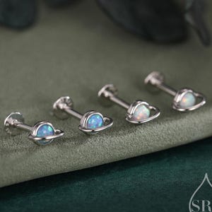 May include: Four silver-toned ear studs with a celestial theme. Two studs feature a planet design with a blue opal center, while the other two have a heart-shaped frame with a white opal center. The studs are displayed on a green surface.