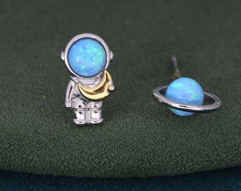 Puede incluir: Un par de pendientes de plata con forma de astronauta y planeta con piedras de ópalo azul. El astronauta sostiene una luna creciente.