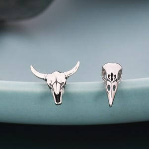 May include: A pair of silver-toned stud earrings. One earring is a bull skull, and the other is a bird skull. The earrings are displayed on a light blue plate with a blurred green leaf in the background.