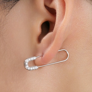 May include: A silver safety pin earring with clear cubic zirconia stones. The earring is worn in the left ear of a person.