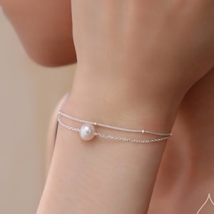 May include: A delicate silver chain bracelet with a single pearl charm. The bracelet is layered with a thinner chain with small beads.