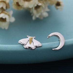 May include: A pair of silver-toned stud earrings. One earring is a crescent moon shape, and the other is a moth with white wings and a yellow body. The earrings are displayed on a light blue surface with white flowers in the background.