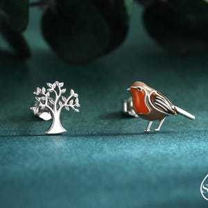 Può includere: Un paio di orecchini a lobo. Uno è un albero d'argento, l'altro un uccellino con petto rosso, dorso marrone e ali bianche e marroni. Gli orecchini sono su uno sfondo verde acqua.