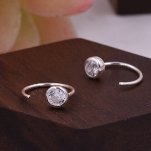 Puede incluir: Dos anillos de nariz de plata con piedras redondas de circonita cúbica transparentes. Los anillos son de estilo aro abierto y están sobre una superficie de madera.