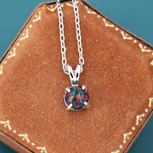 May include: A silver necklace with a round, multi-colored gemstone pendant. The pendant is set in a silver bezel and hangs from a delicate silver chain. The necklace rests on a brown, octagonal jewelry box with gold stitching, against a teal background.