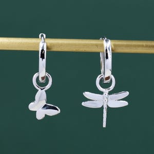 May include: A pair of silver hoop earrings. One earring features a butterfly charm, and the other has a dragonfly charm. The earrings are displayed on a gold-coloured bar against a dark green background.