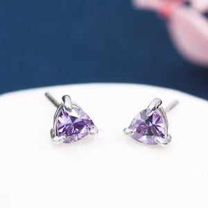 May include: A pair of stud earrings with triangular, light purple gemstones set in silver-coloured metal. The earrings are displayed on a white surface against a dark blue backdrop, with soft pink floral accents in the background.