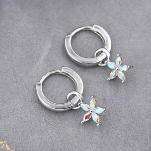 Può includere: Un paio di orecchini a cerchio d'argento con un piccolo ciondolo a forma di fiore che pende da ogni cerchio. I ciondoli a forma di fiore sono fatti di cristalli trasparenti con colori arcobaleno iridescenti.