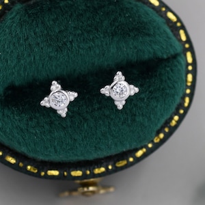 May include: A pair of silver stud earrings with a small round clear stone in the center of each earring. The earrings are surrounded by smaller clear stones. The earrings are in a green velvet box.