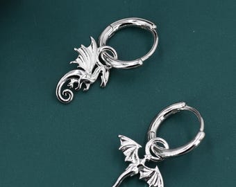 May include: A pair of silver hoop earrings, each with a dragon charm. The dragon charms have detailed wings and a curled tail. The earrings are set against a dark teal background, with a white and black leaf in the upper left corner.