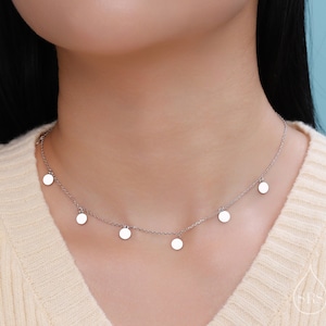 Peut inclure: Un collier en argent avec une chaîne délicate et cinq petits pendentifs ronds. Le collier est porté par une personne avec un pull marron clair.