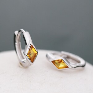 May include: A pair of silver hoop earrings with a small, yellow, diamond-shaped gemstone set in each hoop.