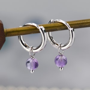 May include: A pair of silver hoop earrings with a small purple gemstone dangling from each hoop.