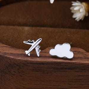May include: A pair of silver stud earrings. One earring is shaped like an airplane, and the other is a cloud. The earrings are displayed on a wooden surface with white flowers in the background. The earrings are small and delicate.