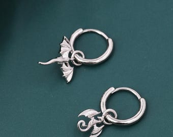 May include: A pair of silver hoop earrings with dragon charms. Each earring has a small, detailed dragon pendant suspended from a circular hoop. The dragon has outstretched wings and a detailed body. The earrings are displayed on a dark teal surface.