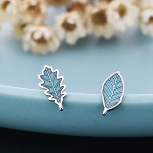 May include: A pair of leaf-shaped earrings with silver edging and light blue enamel. One earring has a jagged edge, the other a smooth edge. They are presented on a light blue surface, with dried flowers in the background.