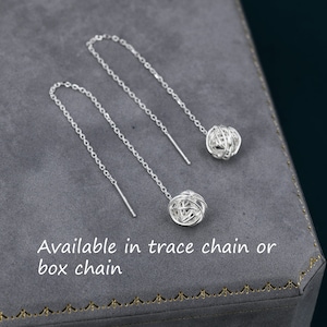 Puede incluir: Un par de pendientes de plata con una pequeña bola redonda envuelta en alambre al final de cada cadena. El texto "Available in trace chain or box chain" es visible debajo de los pendientes.