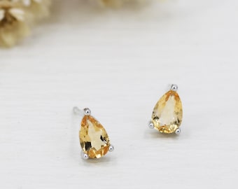 May include: A pair of stud earrings featuring pear-shaped, golden-yellow gemstones set in silver. The earrings are displayed on a white surface with blurred white and yellow flowers in the background. The gemstones have a polished, smooth appearance.