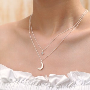 Può includere: Una collana d'argento con un ciondolo a forma di luna crescente e un piccolo ciondolo a forma di stella. La collana è a strati e ha una catena delicata.
